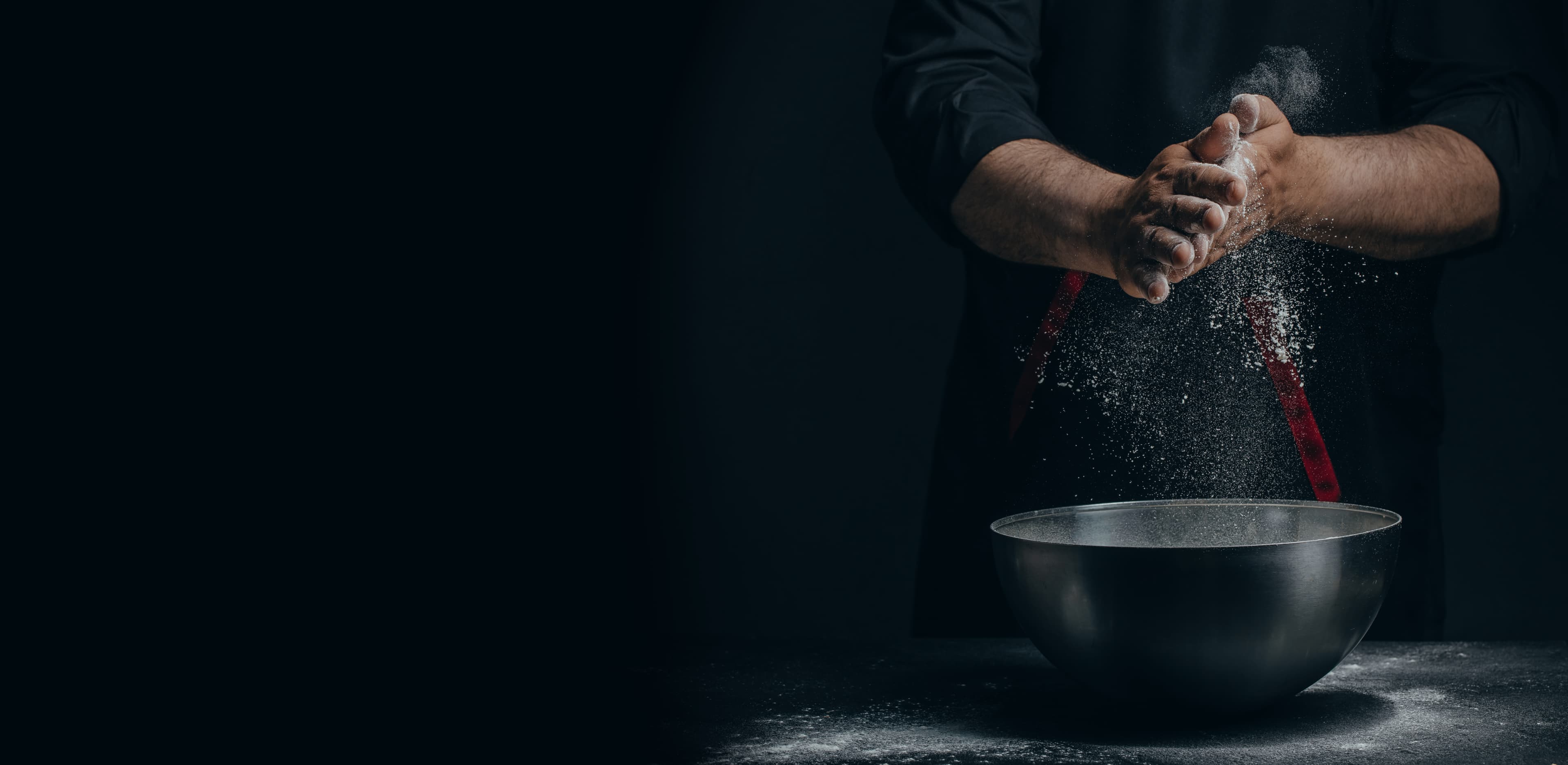 A chef claps flour from their hands above a steel bowl in a dark kitchen.
